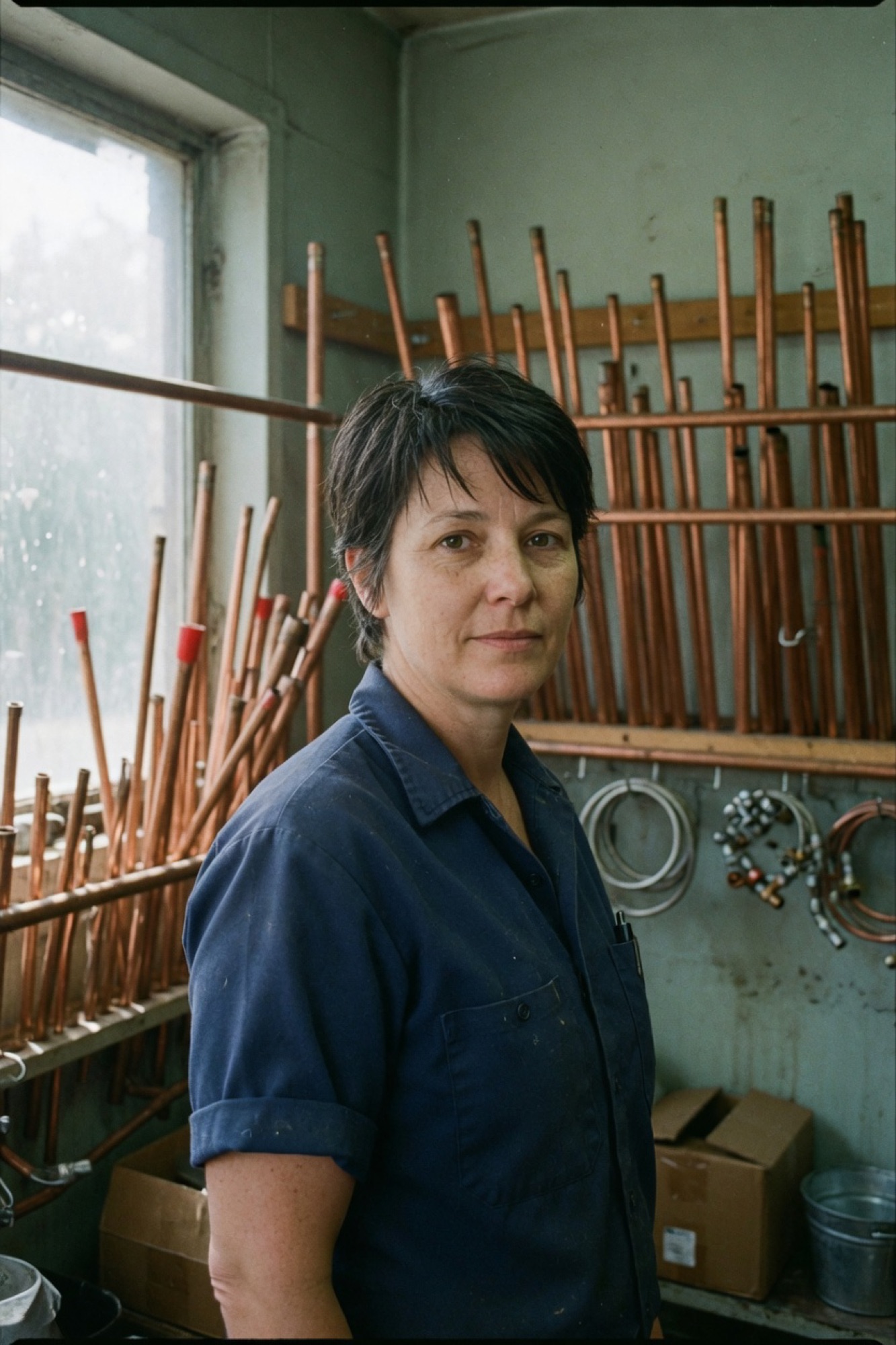 June Calder in the shop, navy work shirt, copper pipe behind her.